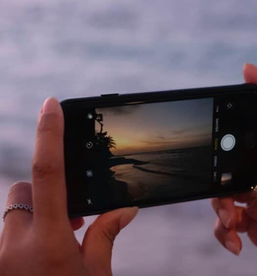 Mãos brancas de uma mulher com alguns anéis. A mulher tira uma foto do pôr do sol na praia. Ao fundo e desfocado está o mar. - Foto: Mackenzie Marco via Unsplash