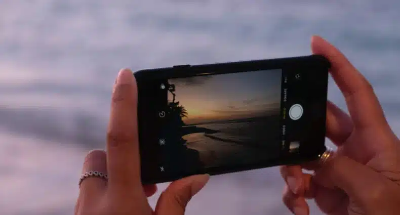 Mãos brancas de uma mulher com alguns anéis. A mulher tira uma foto do pôr do sol na praia. Ao fundo e desfocado está o mar. - Foto: Mackenzie Marco via Unsplash