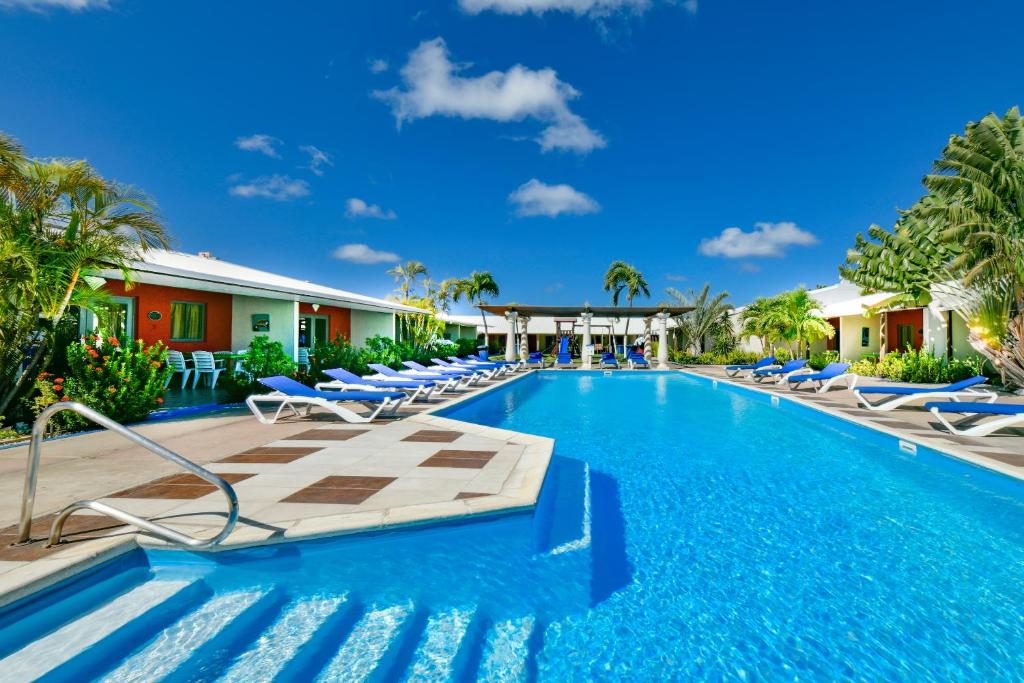 piscina ampla do Aruba Blue Village Hotel and Apartments. Há várias espreguiçadeiras azuis ao redor e diversas palmeiras espalhadas pelo espaço aberto.