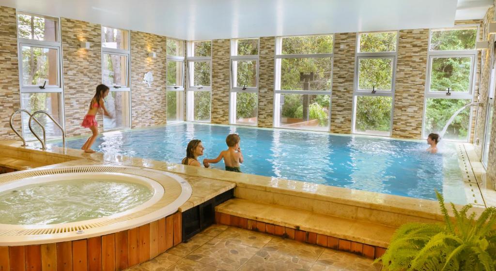 Piscina coberta do Boutique Hotel & Spa Bosque del Nahuel com duas piscinas, uma redonda pequena e uma grande com duas crianças dentro da piscina com um mulher segurando a mão de um menino. Representa hotéis em Bariloche