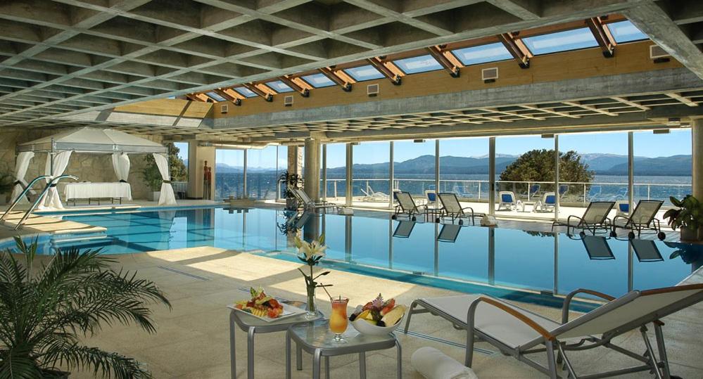Piscina coberta do Cacique Inacayal Lake Hotel & Spa com cadeiras em volta, paredes de vidro com varada e vista para o mar.