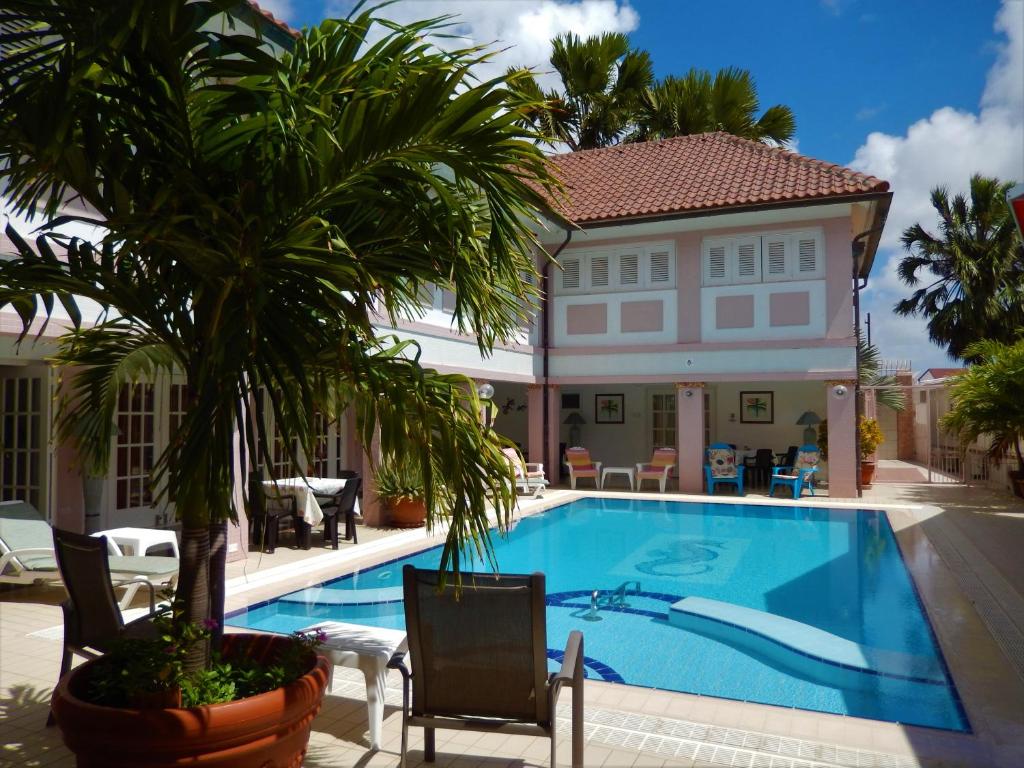 área da piscina do Kamerlingh Villa, um dos hotéis em Aruba, com algumas cadeiras próximas da beirada da piscina e uma varanda coberta e mobiliada ao redor. No canto esquerdo da imagem é possível ver um grande vaso com uma pequena palmeira.