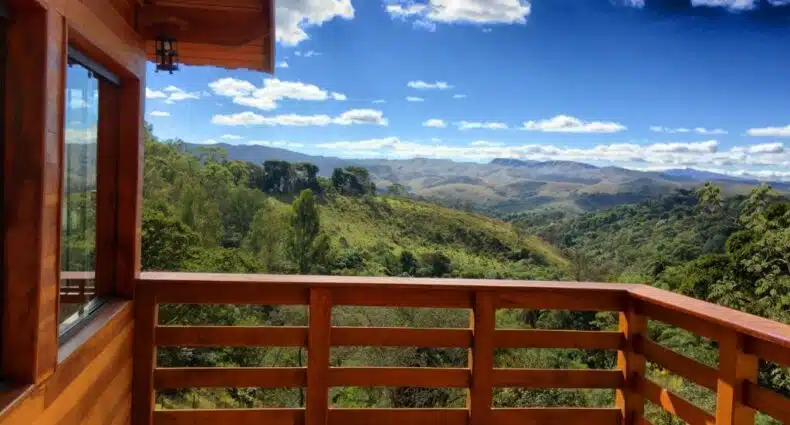 Varanda de um chalé com deck e paredes de madeira, com vista panorâmica da natureza, incluindo montanhas no horizonte, céu azul com nuvens brancas e bastante vegetação verde