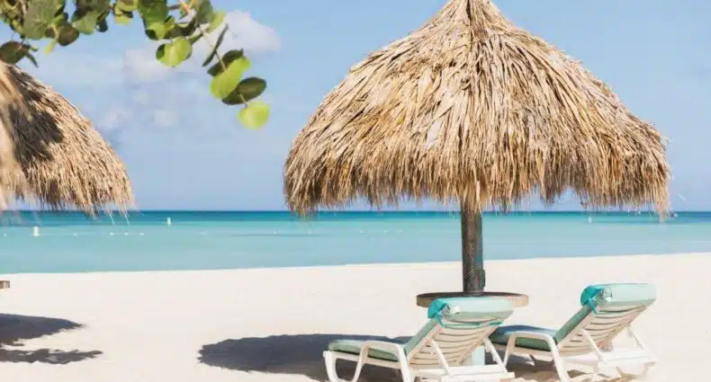 praia em frente ao Boardwalk Boutique Hotel Aruba com duas espreguiçadeiras brancas e estofadas viradas de frente para o mar, sob um guarda-sol de palha
