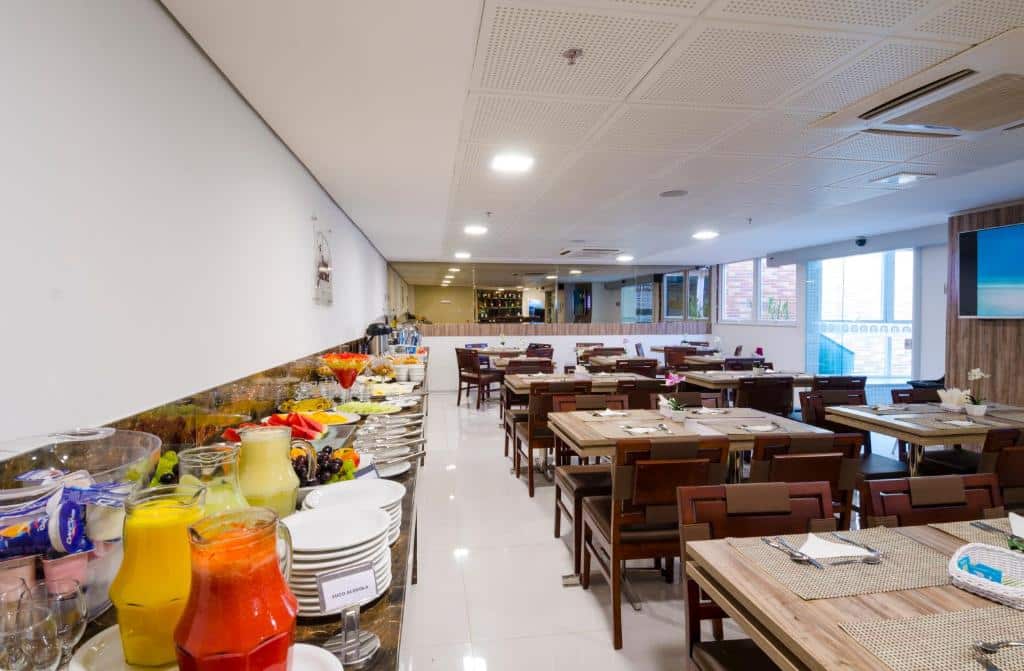 Área de refeições do Profissionalle Hotel São Luís, do lado esquerdo há pratos, sucos, frutas, taças e do lado direito estão as mesas e cadeiras de madeira
