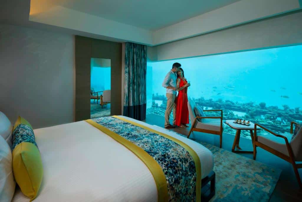 Quarto do Pullman Maldives All-Inclusive, com cama de casal e um casal abraçado ao lado de uma parede de vidro com vista do fundo do oceano. Há também uma mesa pequena com duas cadeiras e um espelho. Imagem para ilustrar o post de hotéis em Maldivas