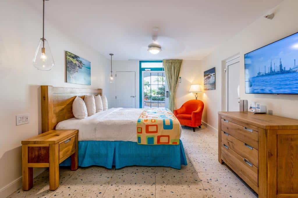 quarto do Amsterdam Manor Beach Resort com uma cama de casal com cabeceira de madeira à esquerda da imagem, uma cômoda de madeira à direita, abaixo de uma televisão de tela plana presa à parede, e uma poltrona laranja no canto do quarto, ao lado da porta.