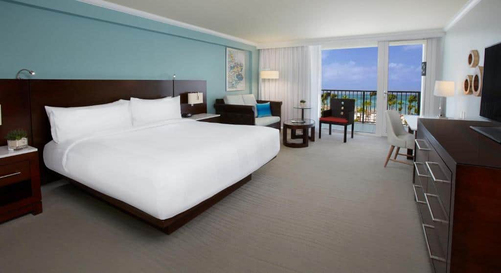 suíte do Aruba Marriott Resort & Stellaris Casino com uma cama de casal à esquerda da imagem, uma área de estar ao lado e uma pequena varanda privativa com vista para a praia e o oceano.