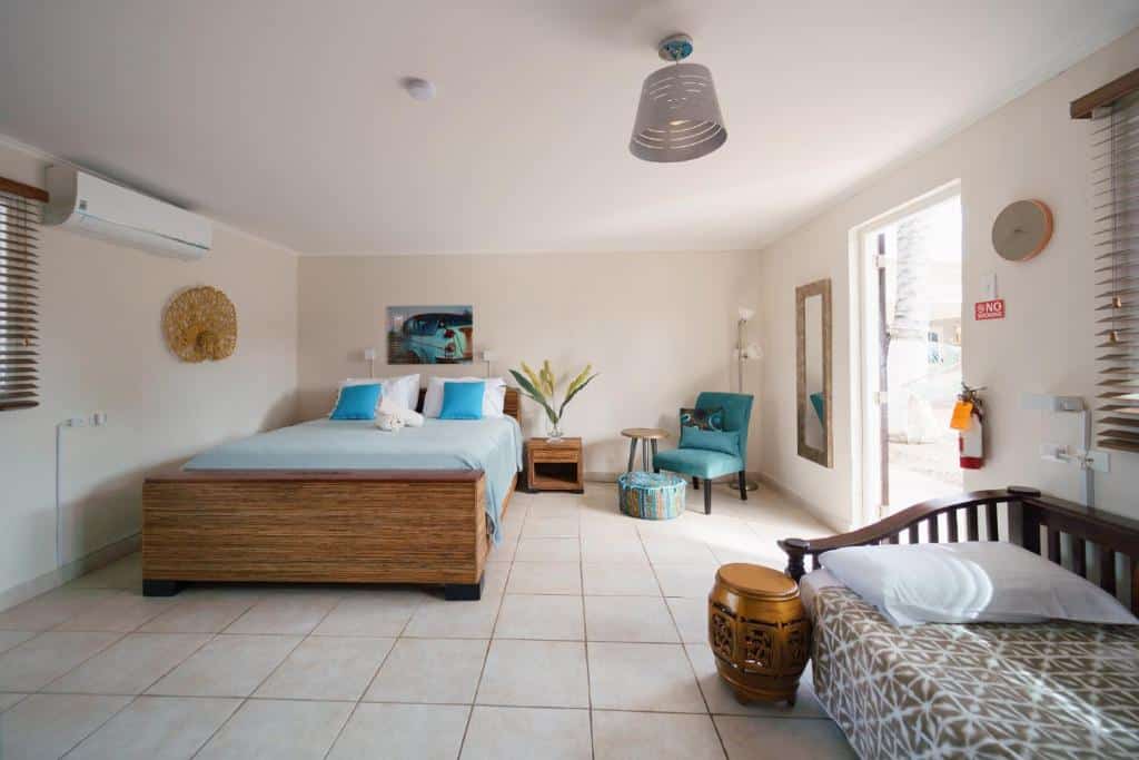 quarto do Arubiana Inn Hotel em Aruba com uma cama de casal, um baú de madeira em frente aos pés da cama, uma pequena poltrona azul no canto direito do quarto, e uma cama de solteiro ao lado da porta que funciona como sofá.