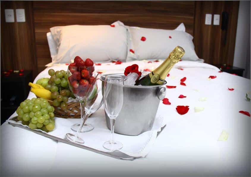 Quarto do Atlântico Golden Apart Hotel com uma cama de casal com pétalas de flores e uma bandeja com um vinho, taças e algumas frutas