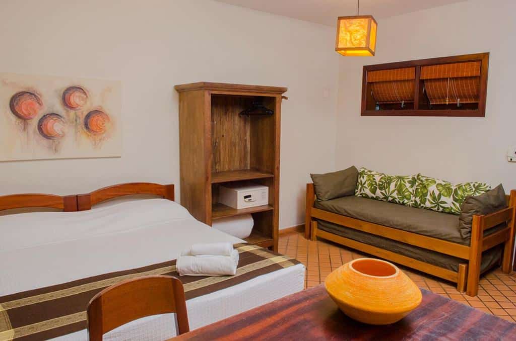 Quarto da Pousada Bico Verde com cama de casal ao centro, cômoda de madeira com cofre, sofá cama do lado direito e, a frente da cama, mesa de trabalho com cadeira.