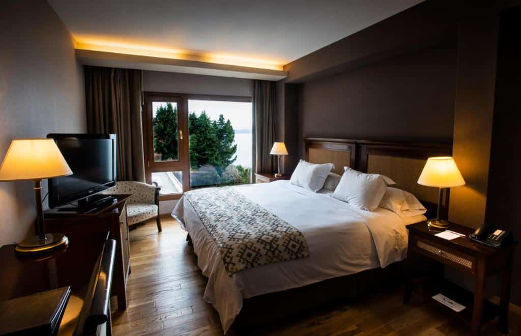 Quarto do Cacique Inacayal Lake Hotel & Spa com cama de casal, duas cômodas com luminárias, cômoda em frente a cama com TV do lado esquerdo poltrona com janelas amplas de madeira e cortinas bege.