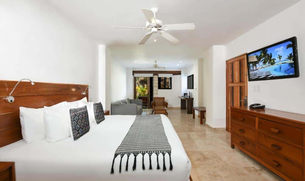 Quarto no Casa del Jaguar Beach Hotel com uma cama de casal, uma televisão e uma cômoda de madeira, no ambiente compartilhado há uma varanda, um sofá, uma poltrona e um banco de madeira