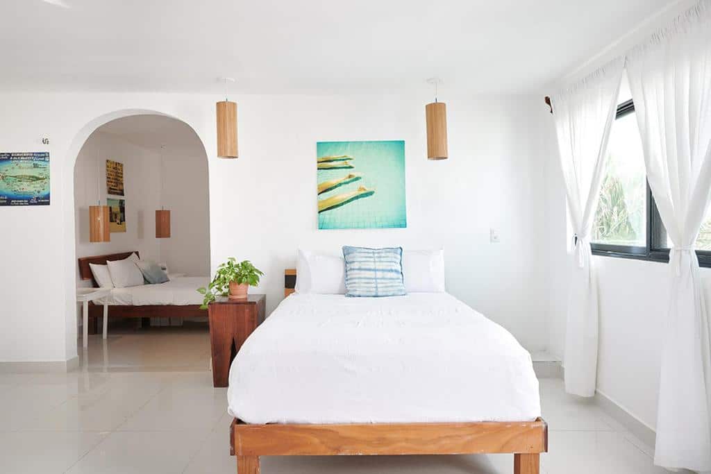 Quarto do Casa el Pio inteiro em branco e com alguns detalhes em madeira, há um cama de solteiro e uma janela com cortinas do lado esquerdo, para representar hotéis em Isla Mujeres