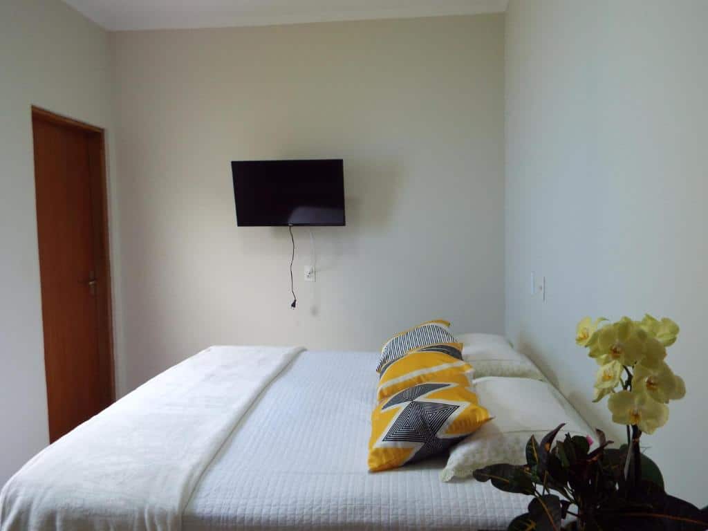 Quarto da Casa Premiatto com cama de casal, um vaso de flores amarelas do lado direto e do lado esquerdo TV pendurada na parede.