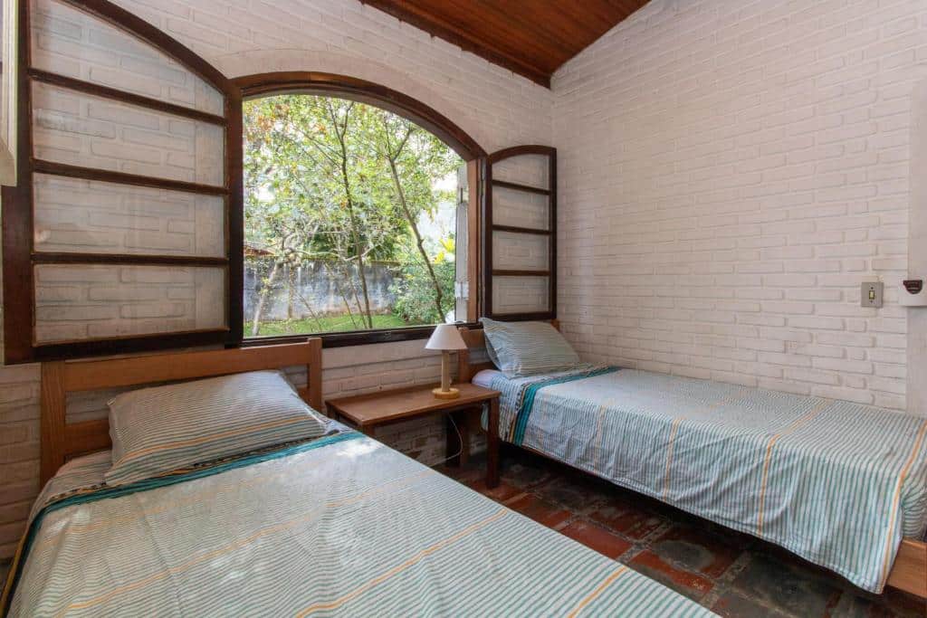 Quarto da Casa Tranquila com duas camas de solteiro e uma cômoda de madeira com luminária e uma janela em cima das camas com vista para o jardim. Representa pousadas na praia do Engenho.