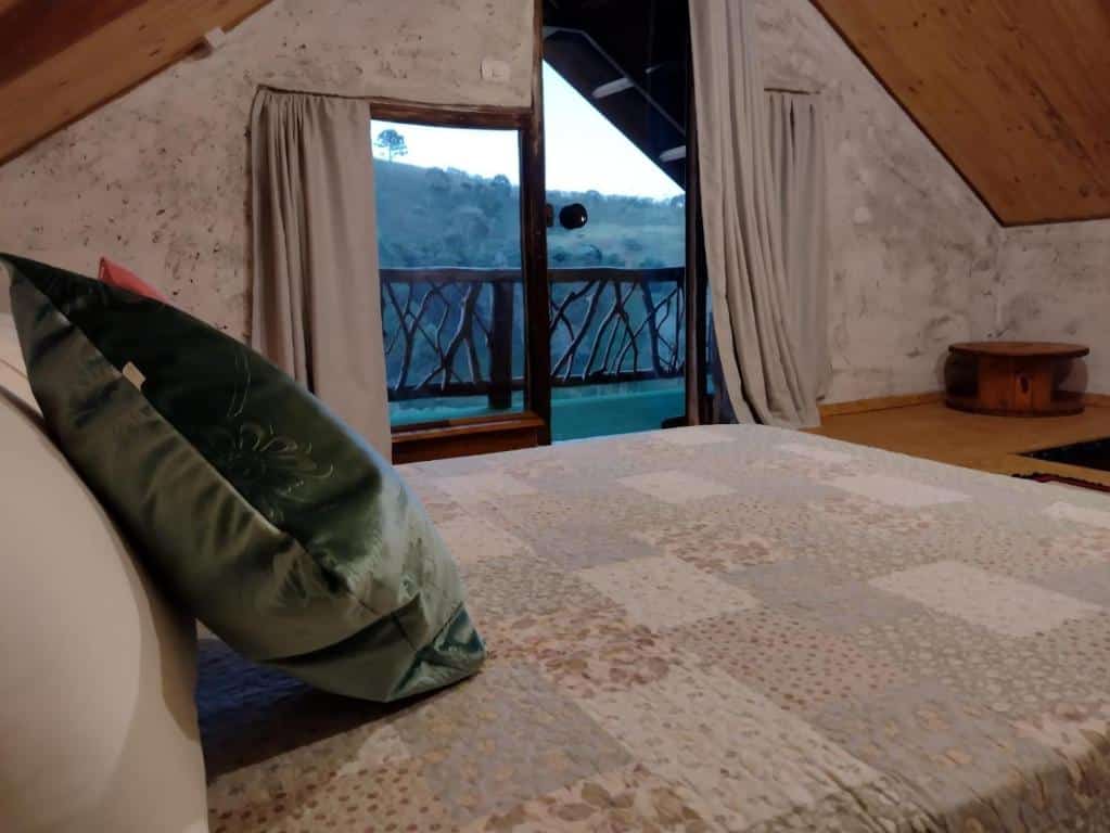 Quarto do Chalés Fenix com cama de casal, cortinas brancas no lado esquerdo com acesso a varanda.