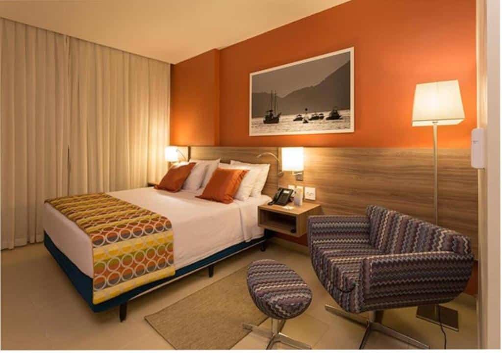 Quarto do Comfort Hotel Santos com uma cama de casal, uma poltrona, um abajur de chão, e uma parede pintada em tom de laranja