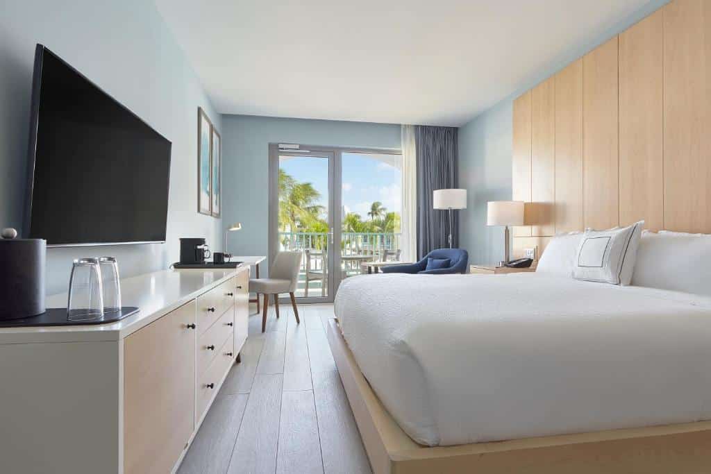 quarto do Courtyard by Marriott Aruba Resort com uma cama de casal à direita, uma televisão de tela plana presa à parede à esquerda da imagem, portas amplas ao fundo com vista para palmeiras e a área comum do hotel