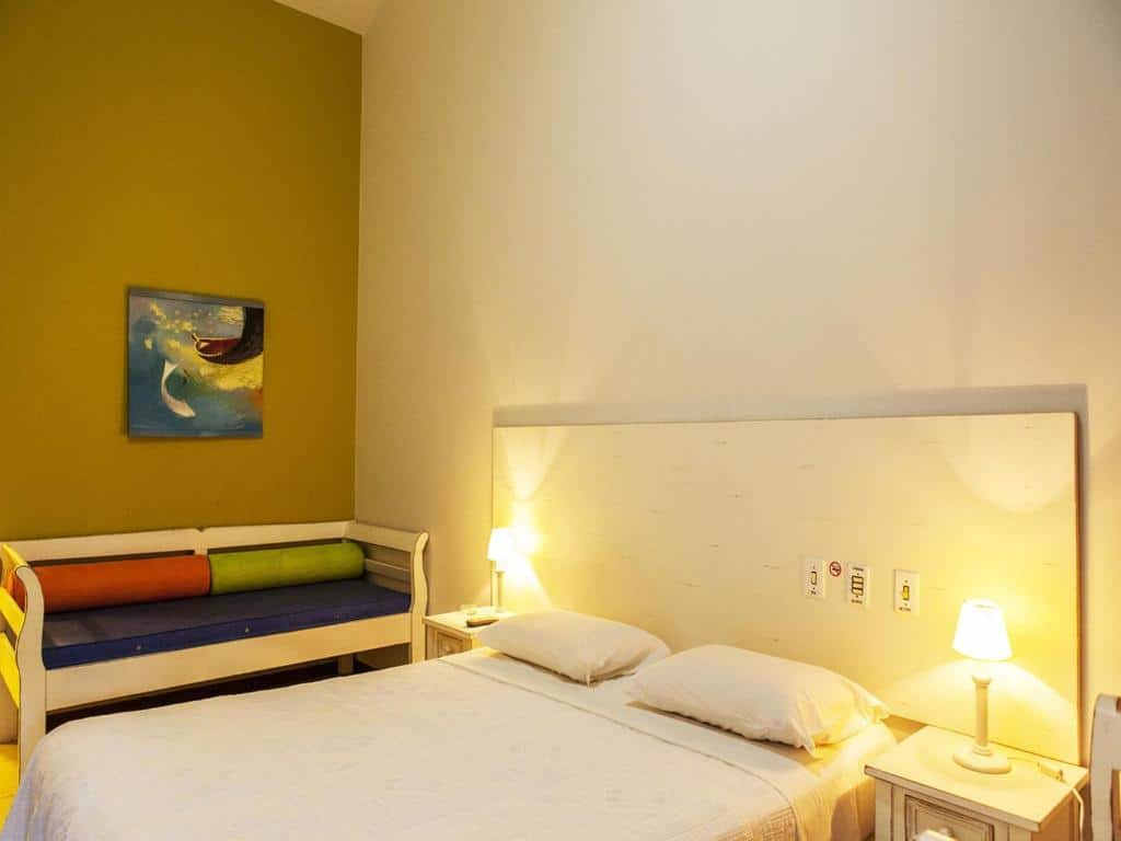 Quarto da Pousada Ponta do Lobo, de 20 m², com uma cama de casal com uma mesinha com abajur aceso de cada lado e um sofá ao lado para ilustrar as pousadas em Balneário Camboriú