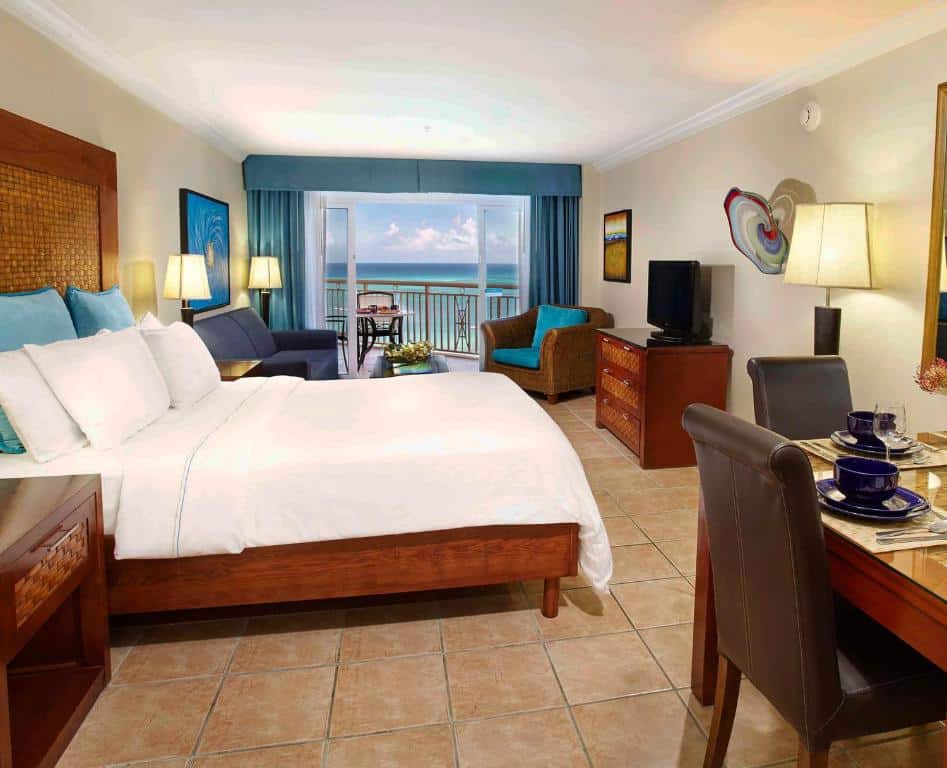 quarto do Divi Aruba Phoenix Beach Resort com uma cama de casal com cabeceira de madeira à esquerda, uma área de estar com sofá e poltrona entre a cama e a varanda privativa, e uma vista panorâmica do mar turquesa ao fundo