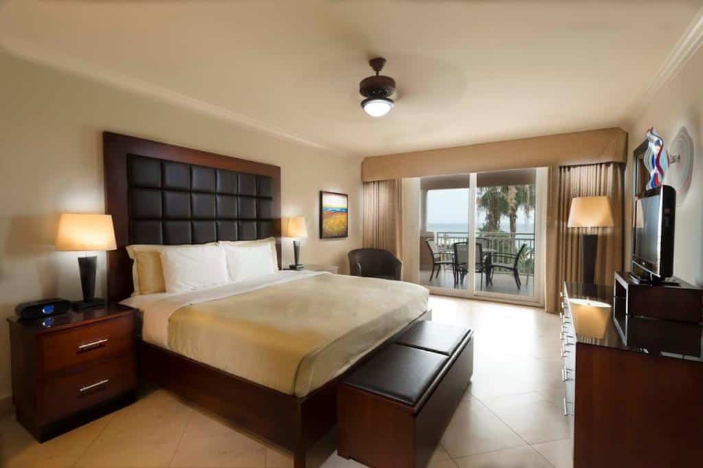 quarto do Divi Village Golf and Beach Resort com varanda em frente ao mar, cama de casal com cabeceira estofada e uma pequena televisão à direita