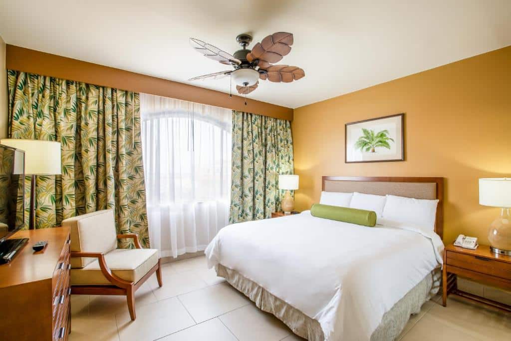 quarto do Eagle Aruba Resort & Casino com um ventilador de telo estilizado, onde as abas são em formato de folhas em madeira. Há uma cama de casal no lado direito e em frente a cama há uma cômoda de madeira com uma televisão em cima.
