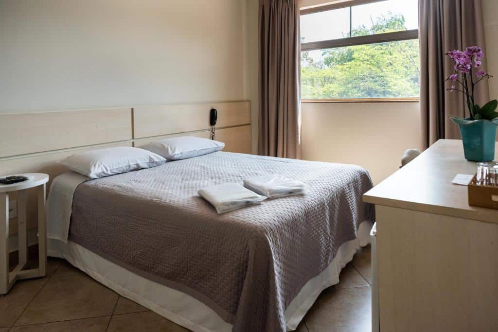 Quarto do Holambra Garden Hotel com cama de casal, uma cômoda do lado direito, janela com cortinas cinzas do lado esquerdo e uma cômoda em frente a cama.