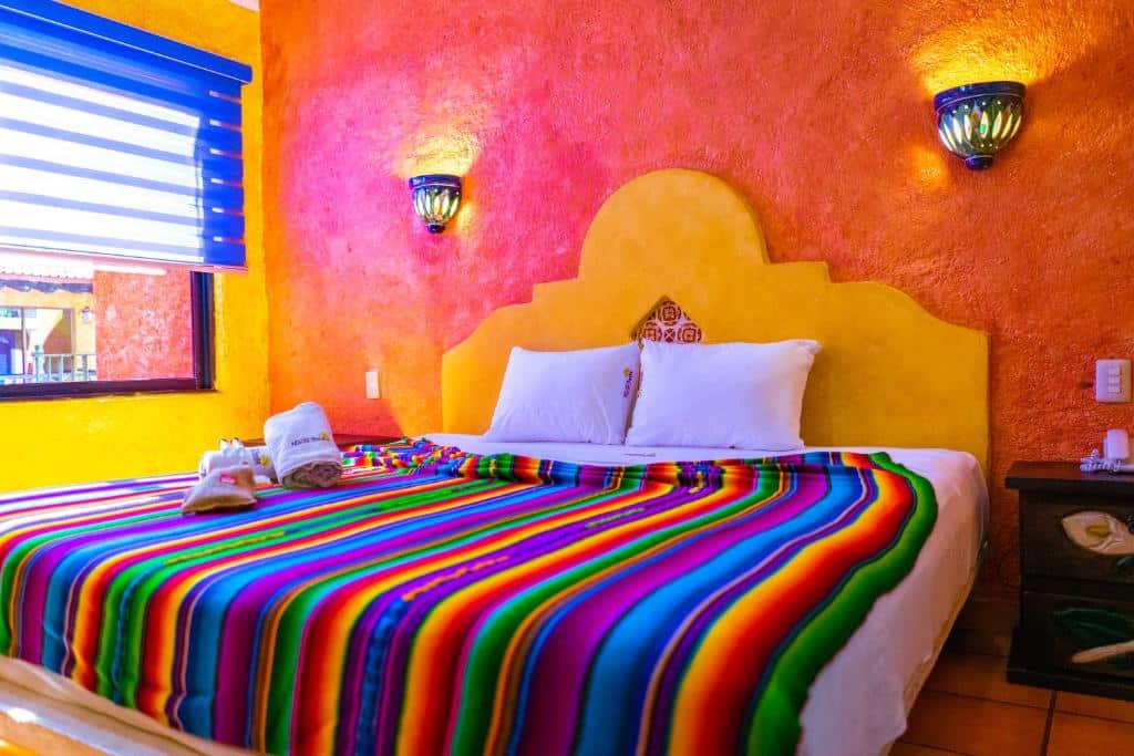 Quarto do Hacienda Maria Bonita Hotel com paredes em tons de laranja, amarelo e rosa, tudo decorado nesses tons, há uma cama de casal e uma janela do lado esquerdo