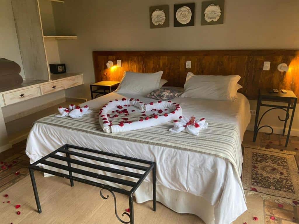 Quarto do Hotel Pousada Shangri-la com cama de casal, duas cômodas de madeira do lado da cama, um banco de ferro na beira da cama, e tolhas com pétalas de rosas vermelhas em cima da cama.