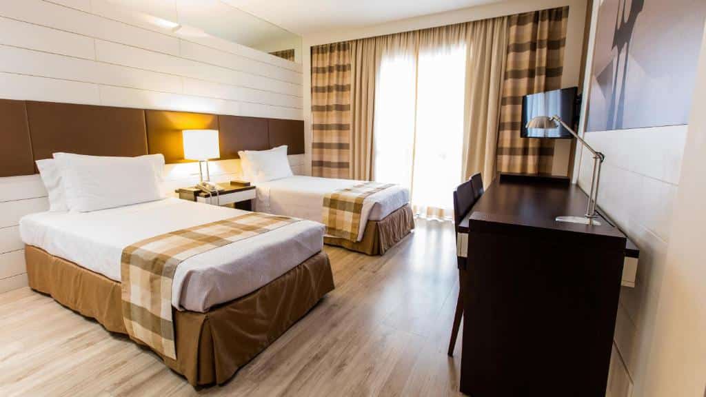 Quarto no Kubitschek Plaza Hotel com duas camas de solteiro, uma varanda com cortinas, uma mesa de escritório de frente com a cama e duas cadeiras, há também uma televisão e o chão é de madeira, para representar os hotéis perto do consulado americano em Brasília