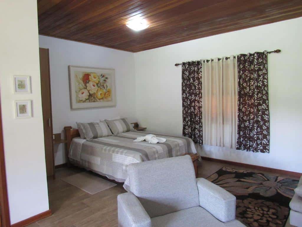 Quarto do Chalés Luar da Serra com cama de casal, toalha branca em cima da cama, cortinas na janela do lado esquerdo e uma poltrona cinza do lado direito.