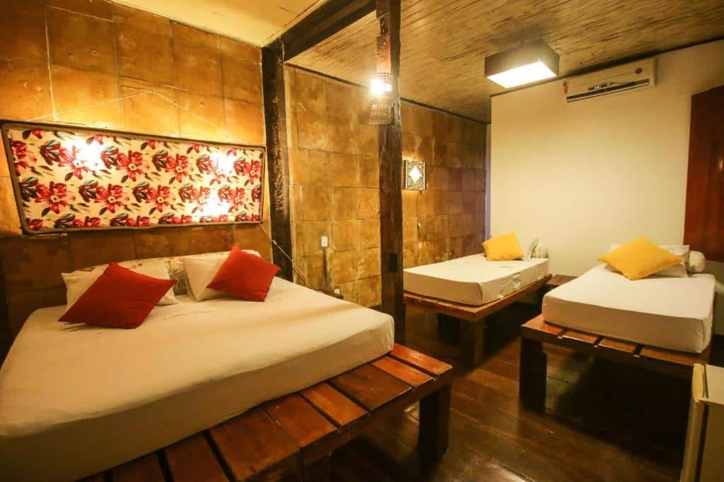 Quarto no Palma Hostel com uma cama de casal e duas de solteiro, em um ambiente rústico com madeira na decoração do espaço e almofadas coloridas sob as camas, para representar hotéis em São Luís do Maranhão
