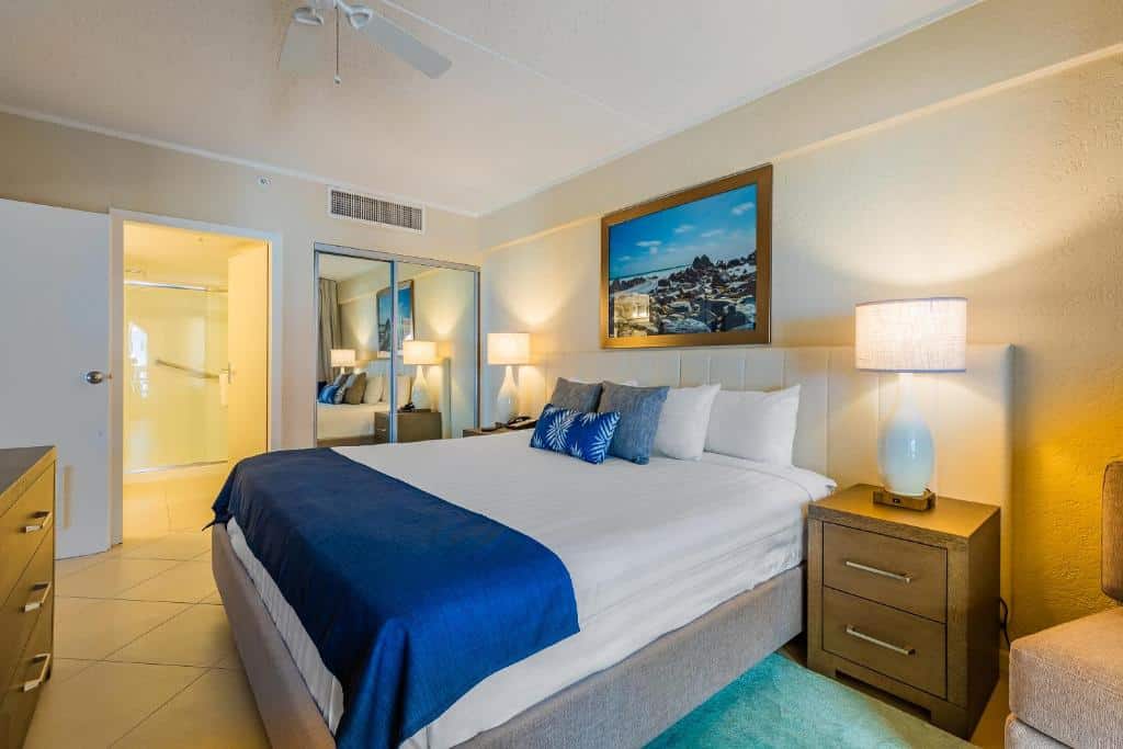 quarto do Playa Linda Beach Resort com uma grande cama de casal no centro do quarto, um quadro da praia na parede em cima da cabeceira estofada e branca da cama. Um banheiro à esquerda e um armário espelhado à direita.