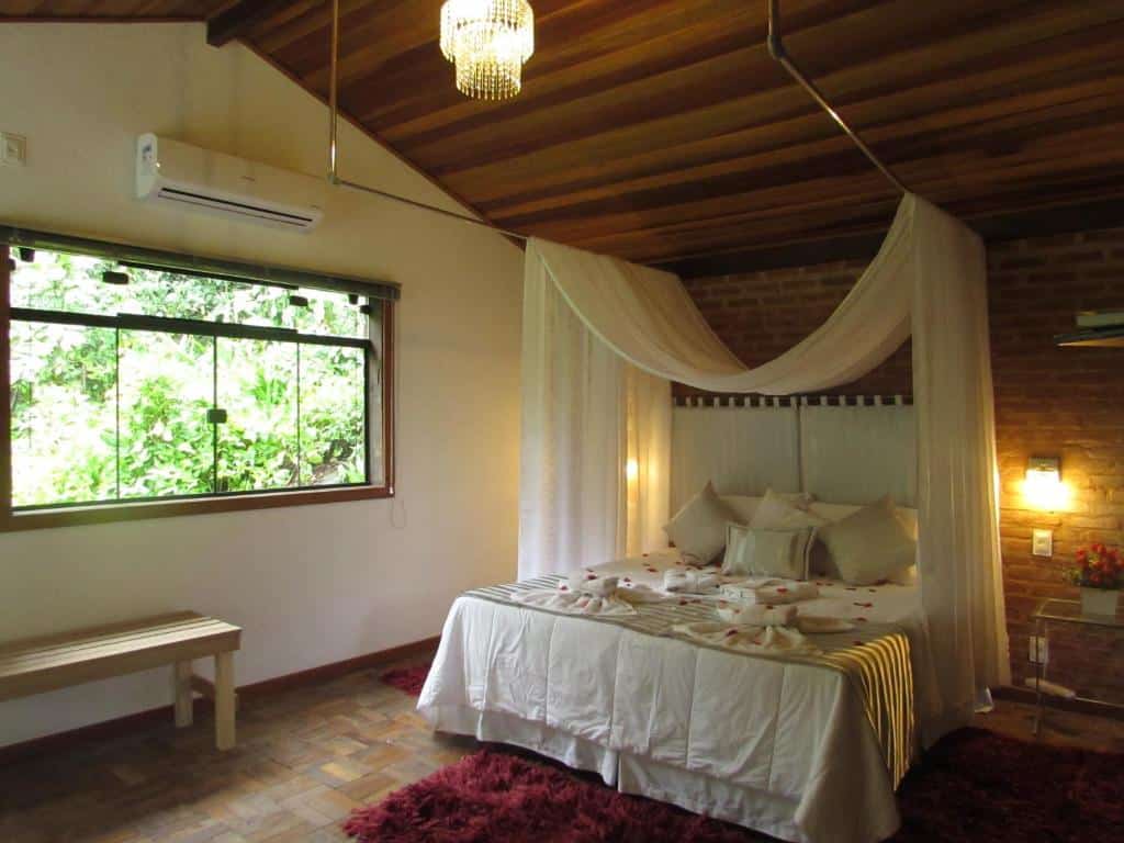 Quarto da pousada Sitio e Poesia em Teresópolis com cama da casal e pétalas de rosa em cima e janela de vidro com vista para as árvores, ilustrando post pousadas em Teresópolis.