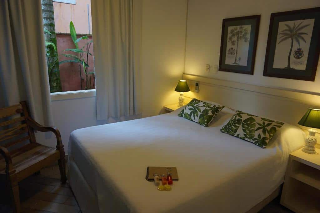 Quarto da  Pousada Terra Juquehy com cama de casal, duas cômodas de ao lado da cama com luminárias, cadeira de madeira do lado esquerdo perto da janela com cortinas brancas.