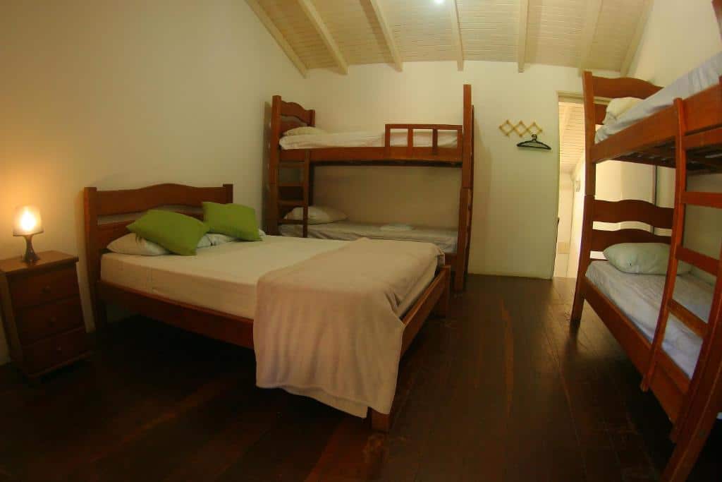 Quarto da Tubes Maresias com cama de casal do lado direito junto a uma cômoda de madeira com luminária, em frente a cama uma cama de beliche e do lado esquerdo outra cama de beliche.