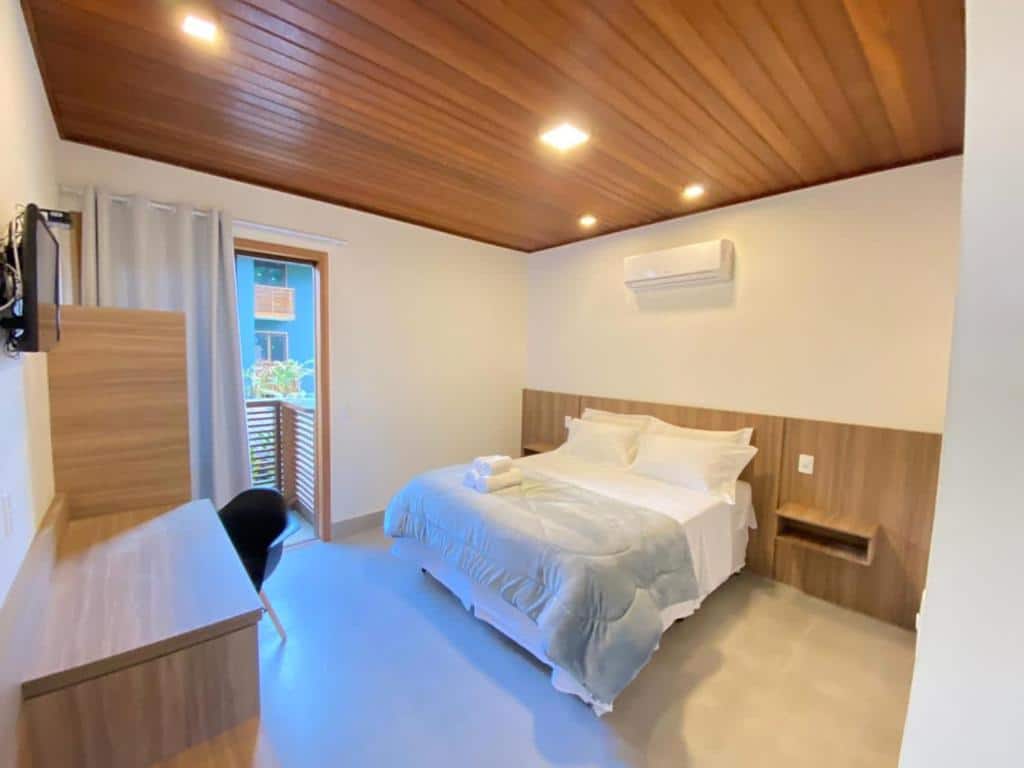 Quarto da Pousada Vila Ipuan com cama de casal no centro do quarto, ar-condicionado a cima da cama, duas cômodas de madeira ao lado da cama, mesa de trabalho em frente a cama e TV acima da mesa de trabalho.