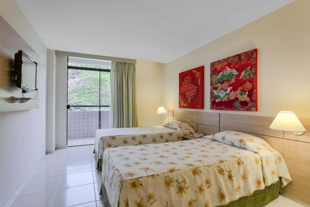 Quarto do Praiabella Hotel com uma pequena varanda com cortinas, duas camas de solteiro com uma cabeceira que tem abajures, sob as camas há dois quadros com pinturas vermelhas e, de frente para as camas, há uma televisão