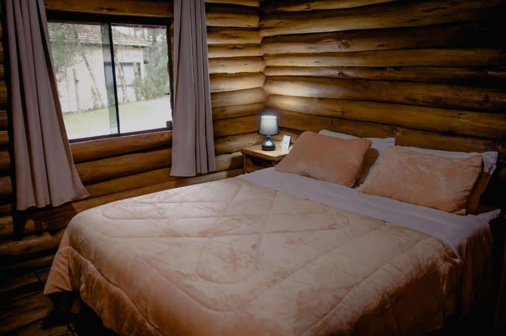 Quarto do Refúgio do Caracol Pipas e Chalés com cama de casal, cômoda do lado esquerdo com luminária perto da janela com cortinas brancas.