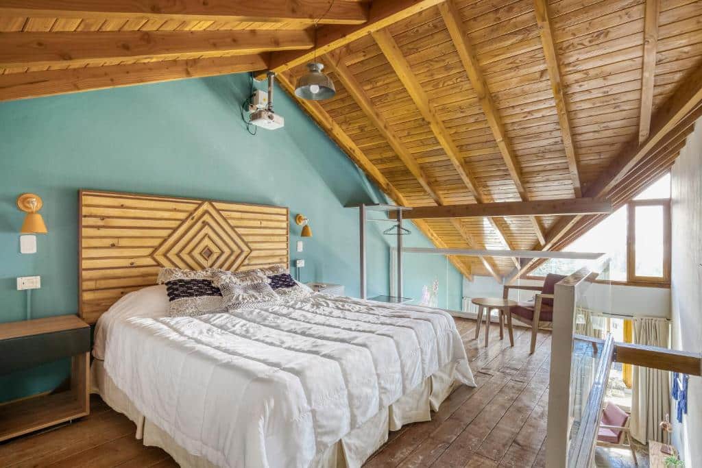 Quarto do Selina Bariloche com cama de casal, duas cômodas de madeira ao lado da cama e uma cadeira com mesinha do lado esquerdo.