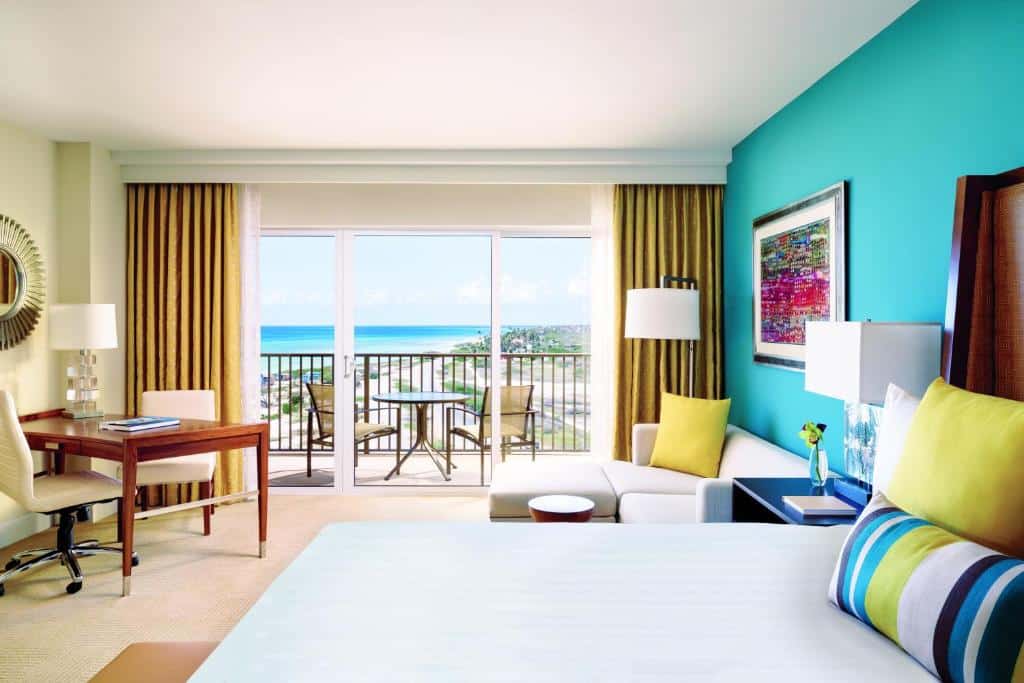 suíte do The Ritz-Carlton, hotel em Aruba, com uma cama de casal e lençóis coloridos, um sofá branco ao lado da cama, uma mesa de madeira no canto esquerdo com uma poltrona branca giratória de frente para as portas amplas de vidro que dão para a sacada privativa. Na sacada há uma mesa redonda pequena e duas cadeiras com vista para a praia e o mar azul turquesa