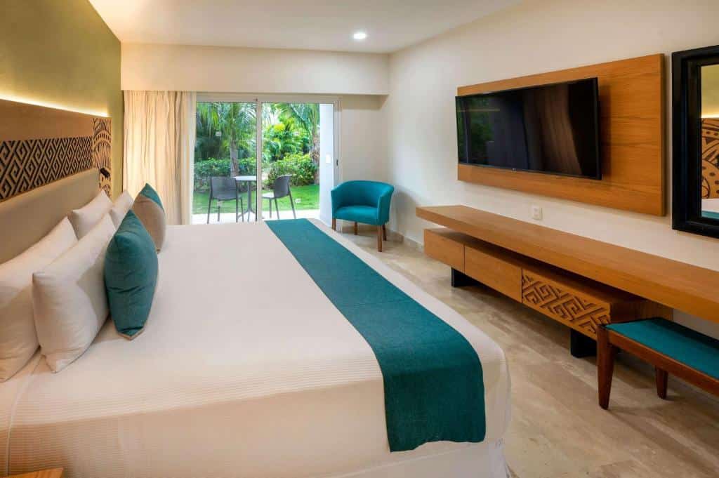 Quarto do Viva Wyndham Azteca All Inclusive com cama de casal, uma varanda com vista pro jardim, uma poltrona estofada em azul, um móvel amplo de madeira e uma televisão de frente para a cama, para representar resorts em Playa del Carmen