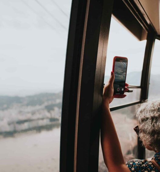 Interior de um teleférico no Rio de Janeiro, com a mão de uma mulher encostada segurando um celular com a câmera tirando foto da paisagem. Há uma vista do mar e casas da cidade no horizonte. Imagem para ilustrar o post de chip celular América do Sul