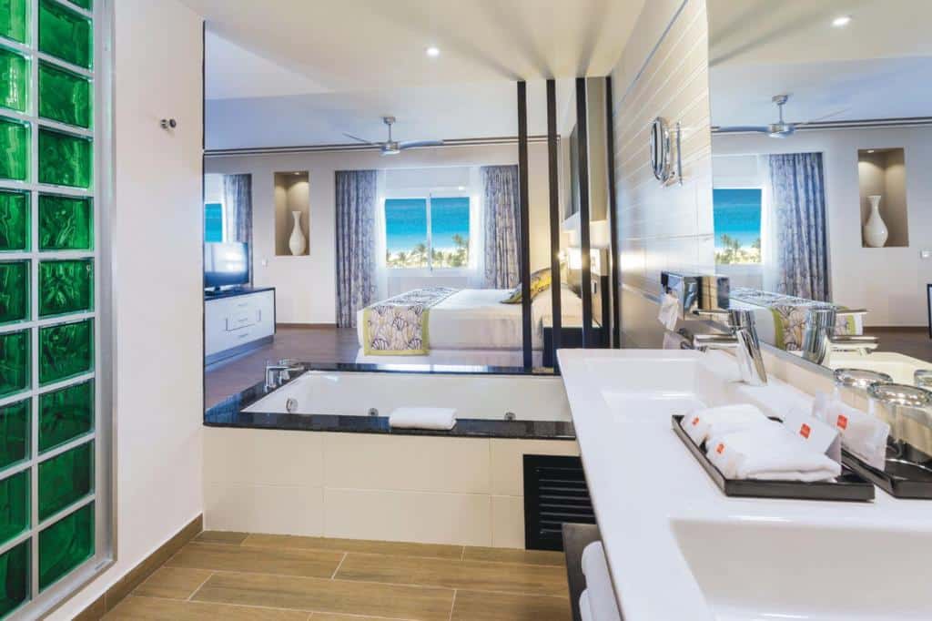 Pia do banheiro do Riu Republica - Adults Only - All Inclusive, com um espelho ao lado refletindo o outro lado do quarto, que tem banheira de hidromassagem, cama de casal, TV e janela com vista do mar azul com árvores na frente
