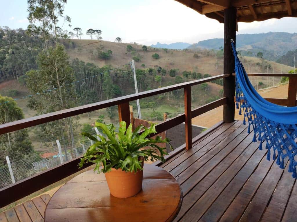 Vista da varanda do Chalés Água azul durante o dia com rede azul do lado esquerdo, mesa e vaso de planta do lado direito e, a frente, tem uma vista para as montanhas e árvores. Representa chalés em Santo Antônio do Pinhal.