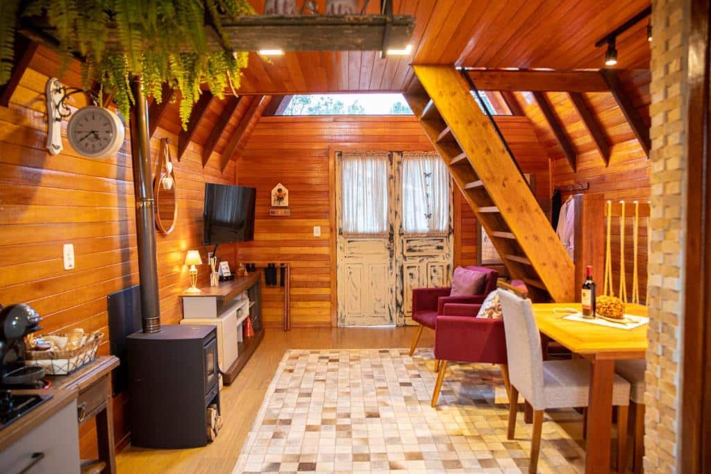 Sala de estar do Cabanas Floratta Wood, com sala no lado direito com TV e cômoda abaixo, duas poltronas cor vinho e uma escada que dá acesso ao quarto, lareira no lado esquerdo com mesa de dois lugares.