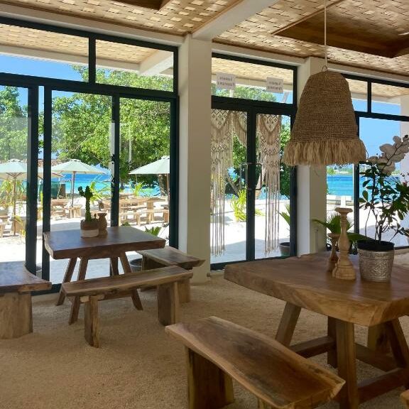 Mesas e cadeiras de madeira num espaço rústico coberto e com portas de vidro mostrando a área beira-mar do Samura Panorama Guest House, que tem mesas e cadeiras de madeira, com guarda-sóis, árvores e mar azul turquesa. Imagem para ilustrar os hotéis nas Maldivas