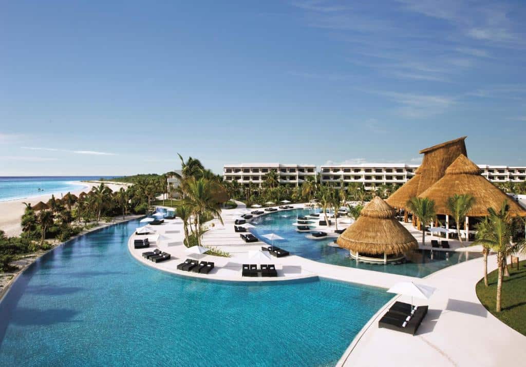 Vista a´rea da um local com diversas piscinas, espreguiçadeiras, coqueiros e o prédio do Secrets Maroma Beach Riviera Cancun - Adults only mais ao fundo, para representar hotéis em Playa del Carmen