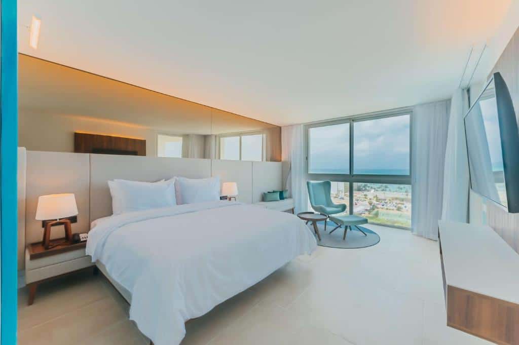 quarto moderno do Radisson Blu Aruba com vista das janelas amplas para a praia. No quarto há uma parte da parede da cama de casal toda espelhada. Entre a cama há dois abajures, e no lado direito há uma poltrona branca com descanso para os pés.